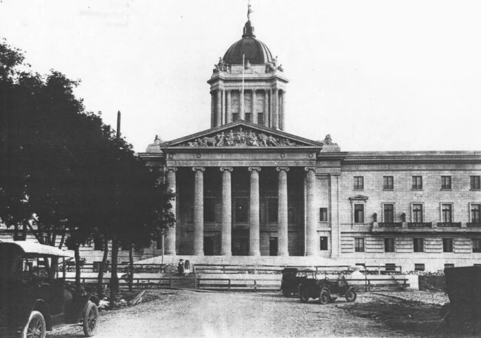 The front facade of the Manitoba Legislative Building with antique vehicles and two people in front.