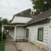 Image 5: A close view of the exterior of the Seven Oaks House from the northeast perspective featuring white slatted exterior and green framed windows