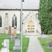 Image 3: Path leading to steps up to large wooden doors with iron detailing that leads into the building with grave markings in view on the grass in front of the building