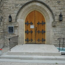 Image 6: Steps up to a large dark stained wooden doors with iron detailings that lead into the cathedral with two wall lamps flanking each side