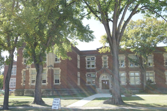 Daniel McIntyre School with trees in front. A sign at the end of the sidewalk reads “PARENT PICK-UP DROP-OFF ZONE.”