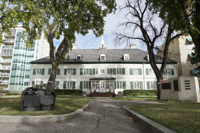 St. Boniface Museum with a hedge and two trees growing in front. Signage sits in the foreground.