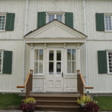 Image 1: Facade of the St. Boniface Museum featuring white slatted exterior, green shutters and a pediment above the front door