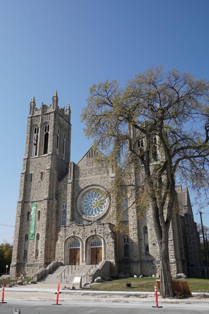 Westminster Church's façade which is made of rough-cut limestone with two double wooden doors in archways atop of steps. Above the doors, there is a large round window. Flanking the doors and window are towers topped with pinnacles.