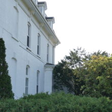 Image 3: A portion of the facade of Government House featuring the white exterior walls, arched windows lining both the first and second floors and greenery around the building