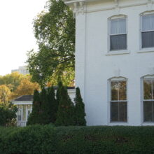 Image 7: A portion of the facade of Government House featuring the white exterior walls, arched windows lining both the first and second floors with a dormer window on the roof and greenery around the building