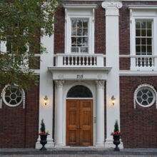 Image 1: Front door of Moss House featuring columns flanking the entrance and 2 round glass paned windows on each side and rectangular windows on the second floor on the red brick facade