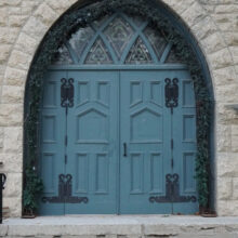 Image 9: Close up view of the blue-green wooden front door of St. Luke's Church featuring coloured glass paned windows above