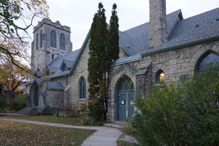 Church tower to the left of a pointed arched two door main entrance on front-facing facade of stone church