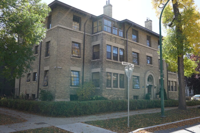 The Wardlaw Apartments behind a hedge. The street sign in front reads “WARDLAW AVE” and “NASSAU ST N.”