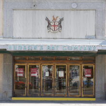 Image 4: One of the entrances to the building featuring brass-coloured doors with glass panes, with the Hudsopn's Bay Company emblem above a sign that reads "Hudson's Bay Company"