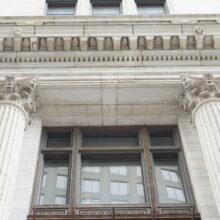 Image 2: Looking up the facade featuring a central large window with two columns flanking featuring decorative bases on them and as well as cornices in view