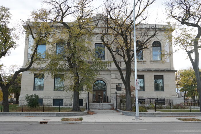 Winnipeg’s Carnegie Library as seen from across the street. Trees frame the entrance and obscure part of the facade.