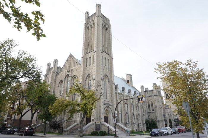 Knox Church with trees and a crosswalk in front of it. A person walks by on the sidewalk.