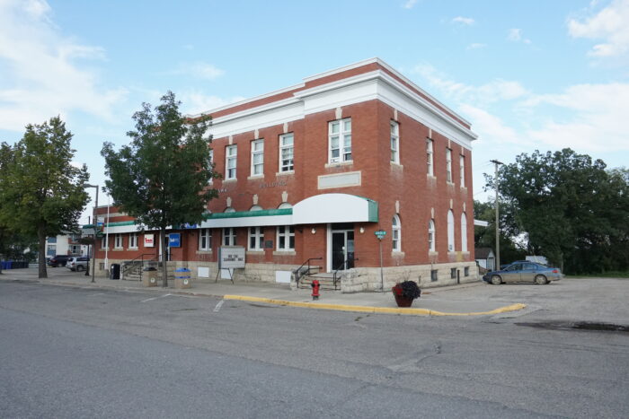 The Neepawa Post Office with “Neepawa Building” displayed on the front facade. A tree grows in front of it.