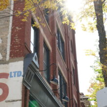 Image 2: The Campbell & Campbell Building with advertising painted on the side. The sun shines through a tree in front.