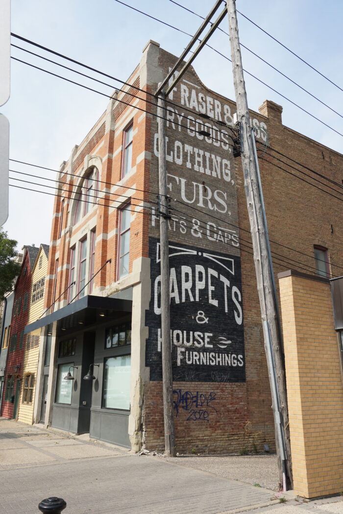 The Fraser Block with advertising painted on the side. Power lines on wooden poles run alongside the building.