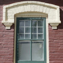 Image 4: Green framed, glass paned window on the facade of the house featuring decorative moulding in ma light colour against the red brick facade