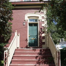 Image 1: Steps up to a red brick house featuring light wooden railings, a green front door, and small glass paned windows with decorative moulding above the door with a dormer window on the roof in view