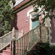 Image 2: Steps up to a red brick house featuring light wooden railings, a green front door, and small glass paned windows with decorative moulding above the door viewed from an angle