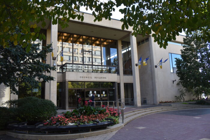 The front facade of Winnipeg’s City Hall Council Building with flags mounted along it. A person walks by.