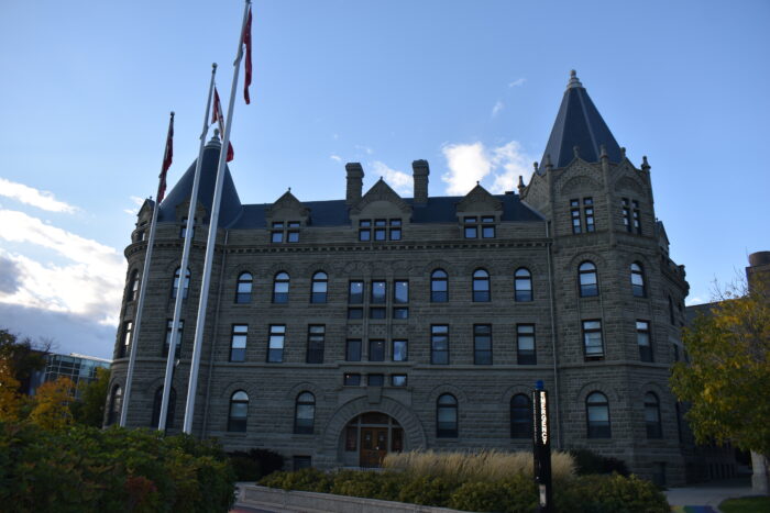 Wesley Hall at the University of Winnipeg with three flag poles and a pillar reading “EMERGENCY” in front.