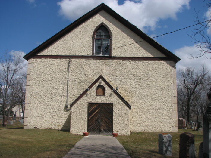 Kildonan Church with a sidewalk leading up to the doors. Headstones are visible in the background and foreground.