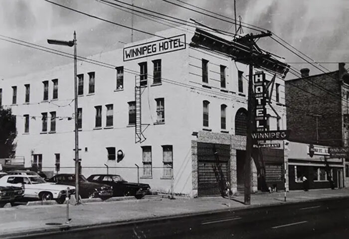 The Winnipeg Hotel with signs reading “WINNIPEG HOTEL” on the front and side. Vehicles parked beside it.