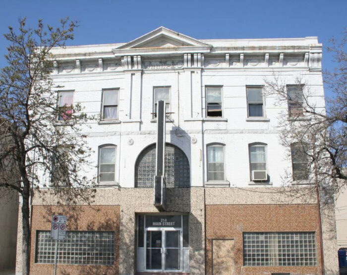 The façade of the Winnipeg hotel in 2020 features double-hung windows, some with segmented arches, and others using glass blocks. At the top of the building, there is a cornice and pediment supported by brackets.